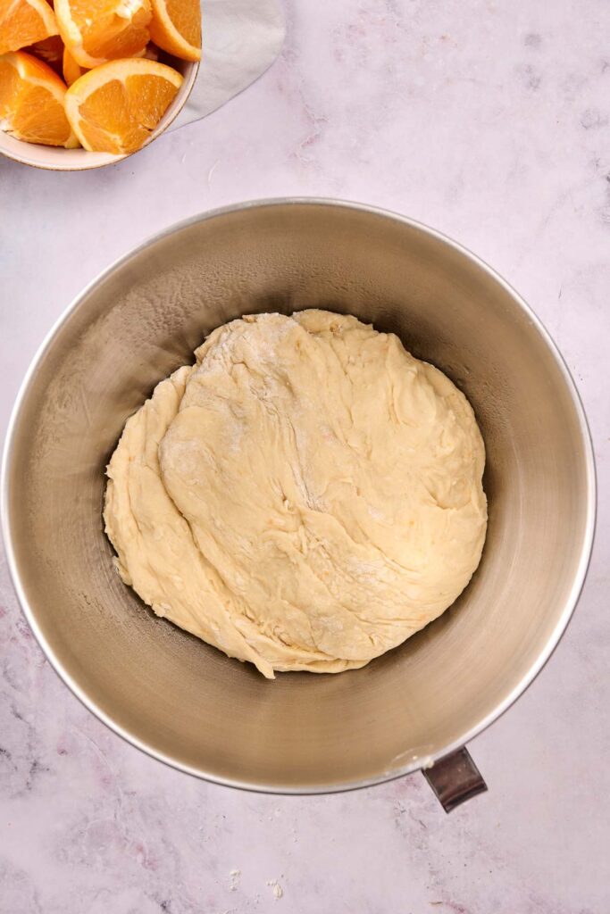A large metal mixing bowl containing a ball of dough for Orange Rolls sits on a light countertop, with a bowl of sliced oranges nearby in the corner.