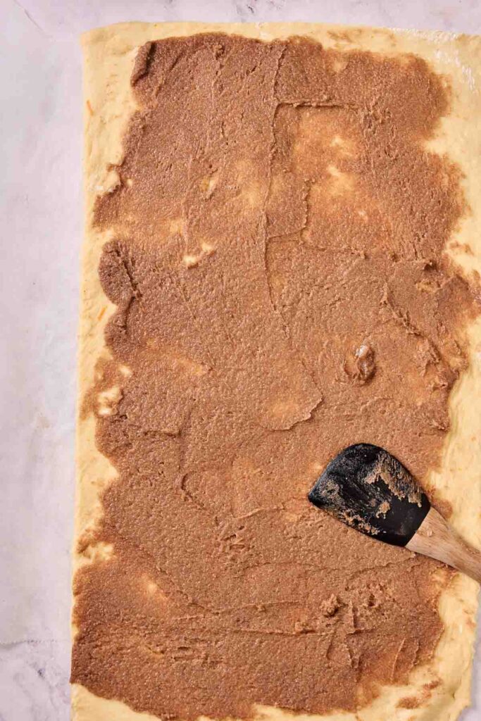 A rectangular sheet of dough spread with a layer of brown cinnamon filling, perfect for making Orange Rolls, with a spatula resting on top.