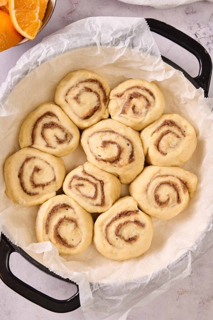 Unbaked Orange Rolls arranged in a parchment-lined round baking dish, ready for baking. Orange slices are partially visible in the corner.
