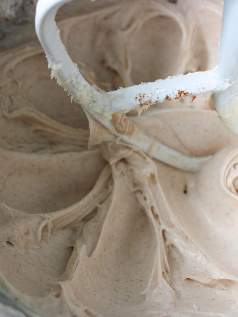 Stand mixer paddle covered in creamy, light brown Pumpkin Cupcakes batter with visible specks, resting above more batter in the mixing bowl.