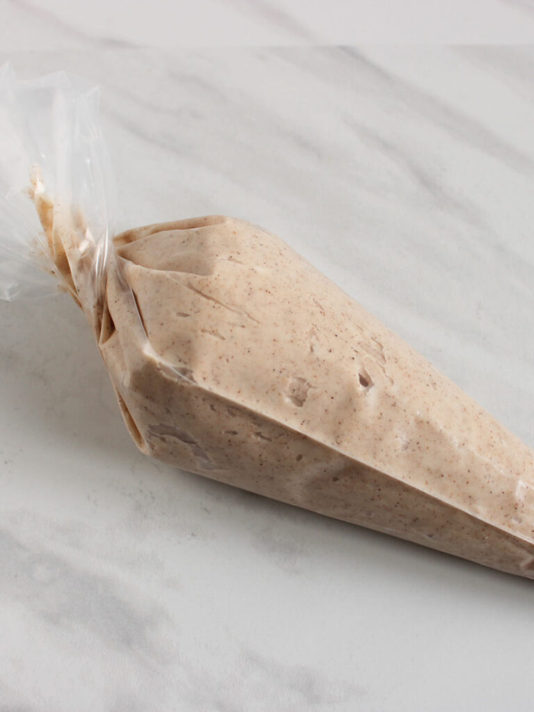 A clear piping bag filled with a light brown, speckled mixture for pumpkin cupcakes, lying on a white marble surface.
