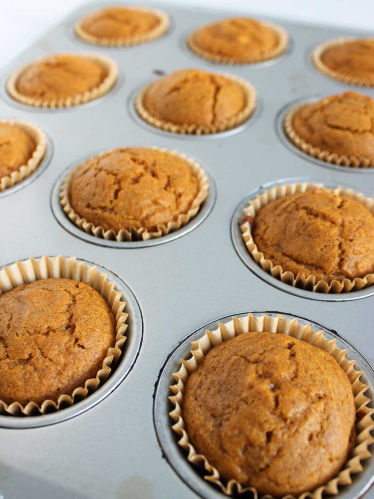 A muffin tin filled with nine freshly baked pumpkin cupcakes in paper liners, their golden brown tops boasting visible cracks.