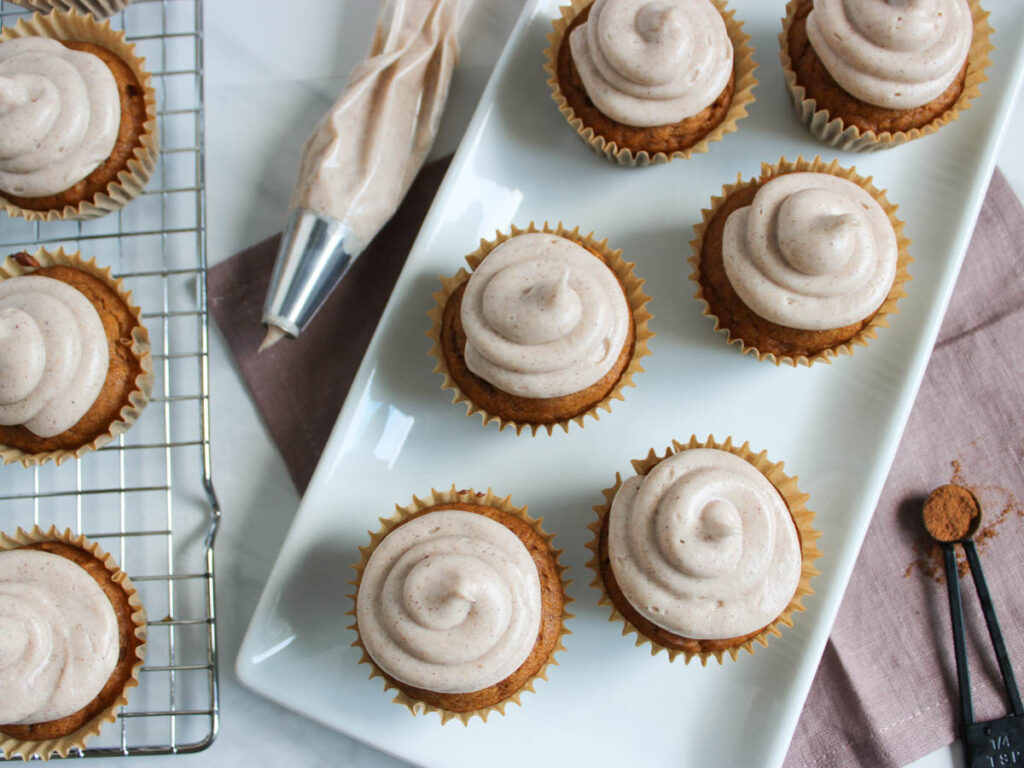 Pumpkin Cupcakes with swirled frosting are arranged on a white plate and cooling rack, beside a piping bag and measuring spoons with cinnamon.