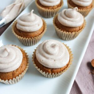 Six Pumpkin Cupcakes with light brown frosting are arranged on a rectangular white plate, with a frosting piping bag visible in the background.