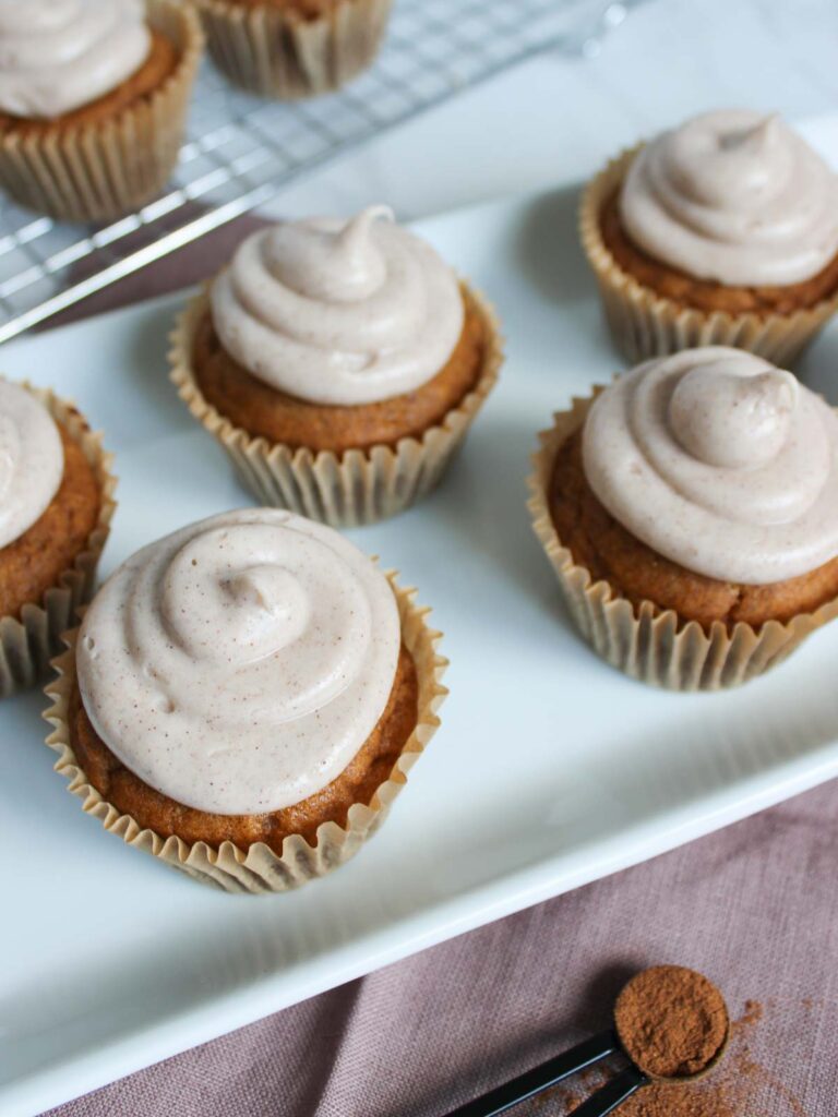 Six pumpkin cupcakes with swirled cinnamon frosting are arranged on a white rectangular plate, with more cupcakes on a cooling rack in the background and a measuring spoon of cinnamon nearby.