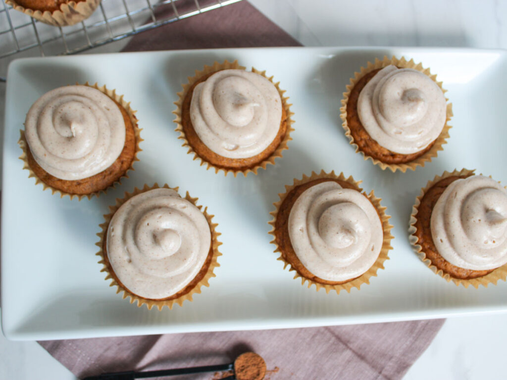 Six Pumpkin Cupcakes with swirled frosting are arranged on a rectangular white plate, with more cupcakes visible on a cooling rack in the background.