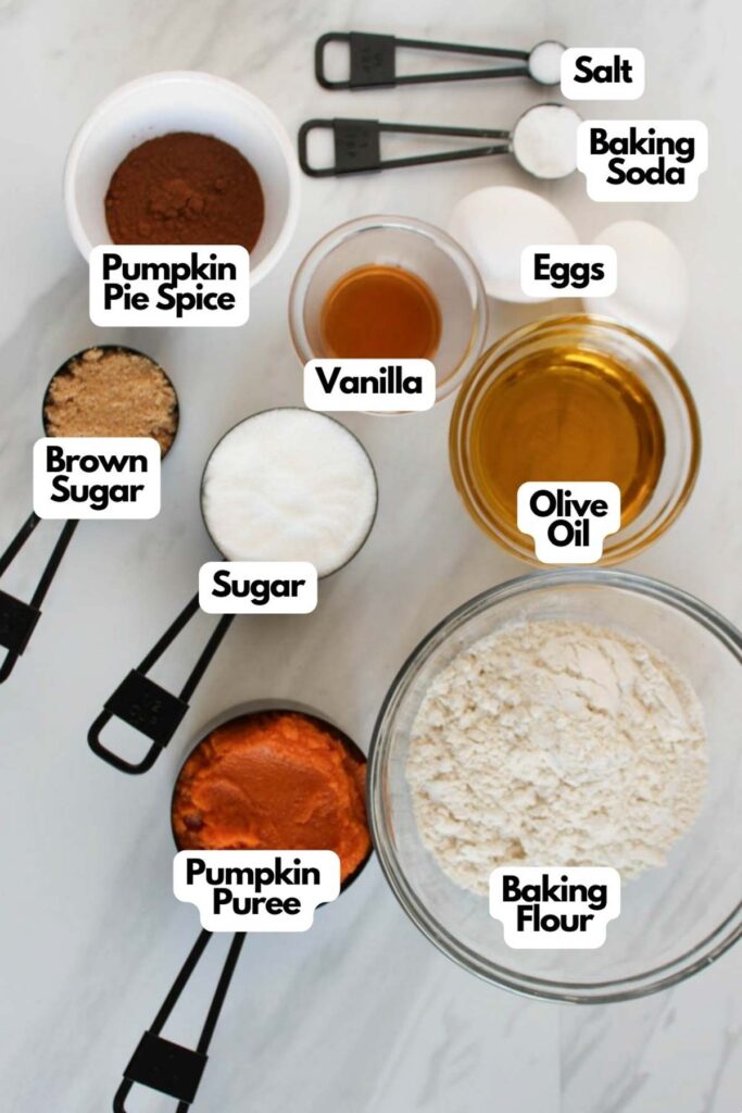 Baking ingredients for Pumpkin Cupcakes displayed on a countertop: pumpkin pie spice, brown sugar, sugar, pumpkin puree, vanilla, eggs, olive oil, baking flour, salt, and baking soda in bowls and measuring cups.