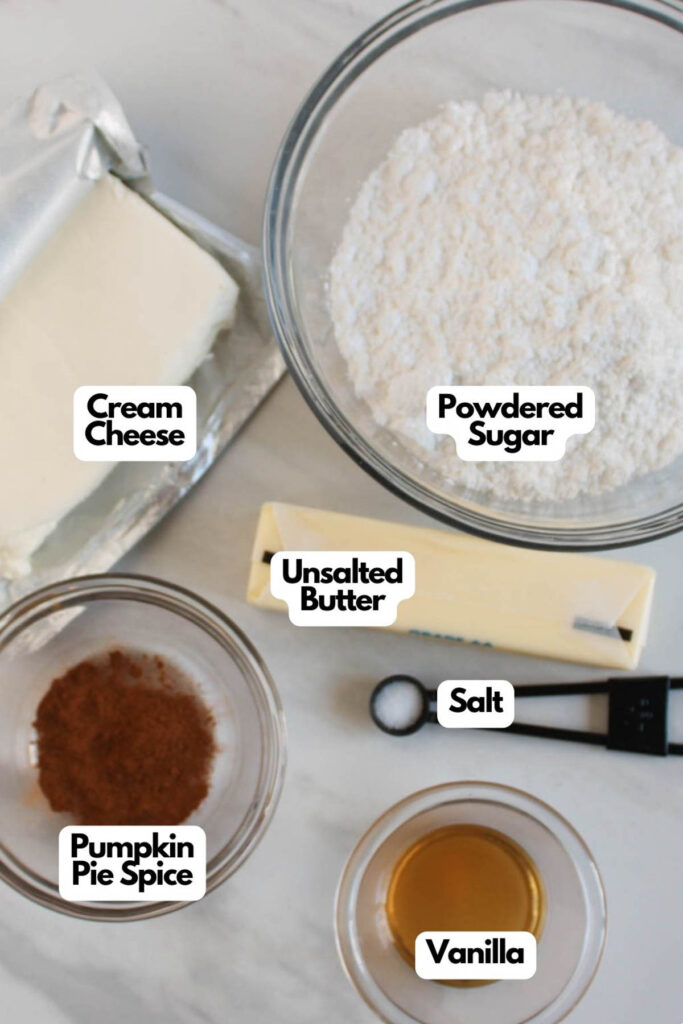 Five labeled ingredients for Pumpkin Cupcakes are arranged on a countertop: cream cheese, powdered sugar, unsalted butter, pumpkin pie spice, vanilla, and salt.