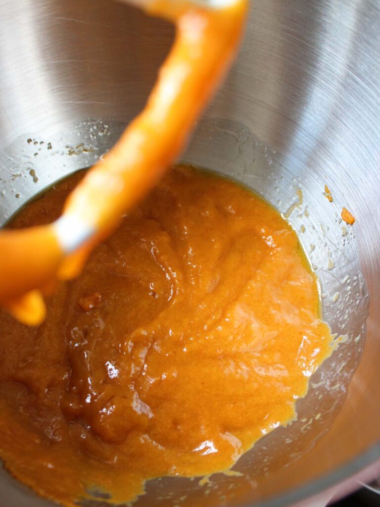 Thick orange batter for Pumpkin Cupcakes being mixed in a stainless steel mixing bowl, with a metal beater partially visible.