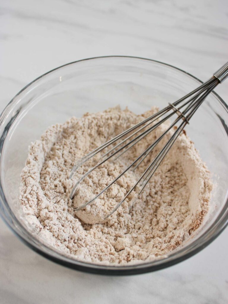A metal whisk rests in a glass bowl filled with flour and other dry ingredients, ready to create delicious pumpkin cupcakes on a white surface.