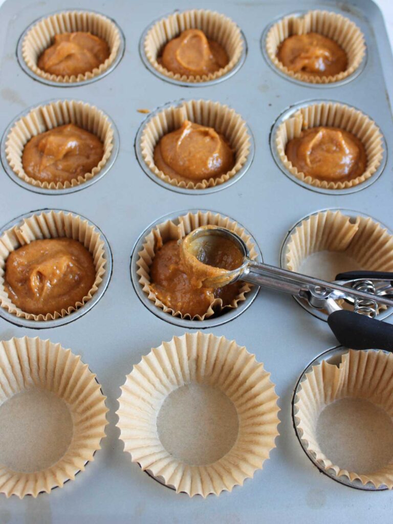 A muffin tin lined with paper cups, some filled with pumpkin cupcake batter and some empty; a scoop with batter hovers above one cup.