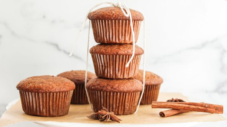 Five muffins arranged on parchment paper, with three stacked and tied with string, next to whole cinnamon sticks and star anise on a white surface.