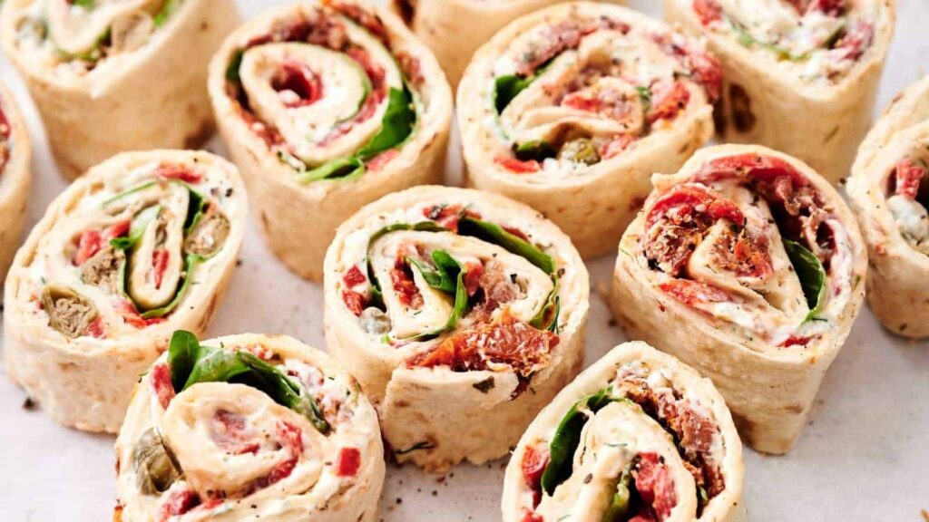 Several tortilla pinwheel sandwiches filled with leafy greens, sliced meats, and spread, arranged closely together on a white surface.