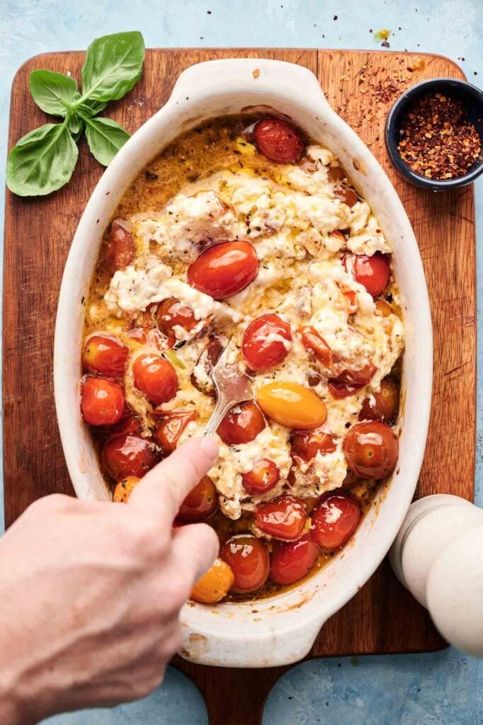 A hand uses a fork to mix baked feta cheese and cherry tomatoes in a white oval dish on a wooden board, creating the classic Baked Feta Pasta. Basil, red pepper flakes, and a pepper grinder add extra flavor nearby.