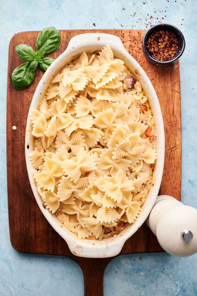 Oval white dish with bowtie pasta atop a creamy Baked Feta Pasta mixture, set on a wooden board with fresh basil and a small bowl of red pepper flakes nearby.
