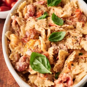 Baked Feta Pasta bowtie noodles with tomatoes, creamy sauce, and fresh basil leaves, served in a white oval dish on a wooden surface.