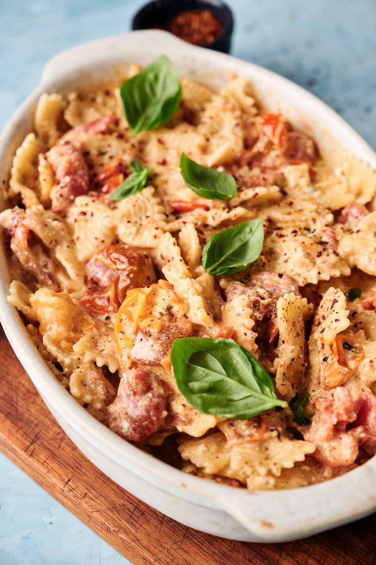 A white dish filled with bowtie pasta in a creamy baked feta pasta sauce, topped with cherry tomatoes and fresh basil leaves, set on a wooden board.