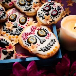 Day of the Dead Cookies decorated with colorful icing and flower accents are arranged in a tray next to a candle and purple flowers, perfectly capturing the festive spirit of this cherished celebration.