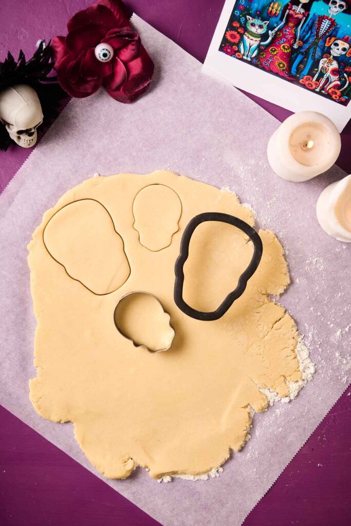 Rolled-out cookie dough on parchment paper with three skull-shaped cutters, surrounded by candles, a skull decoration, and a Day of the Dead-themed photo&mdash;perfect for creating festive Day of the Dead Cookies.