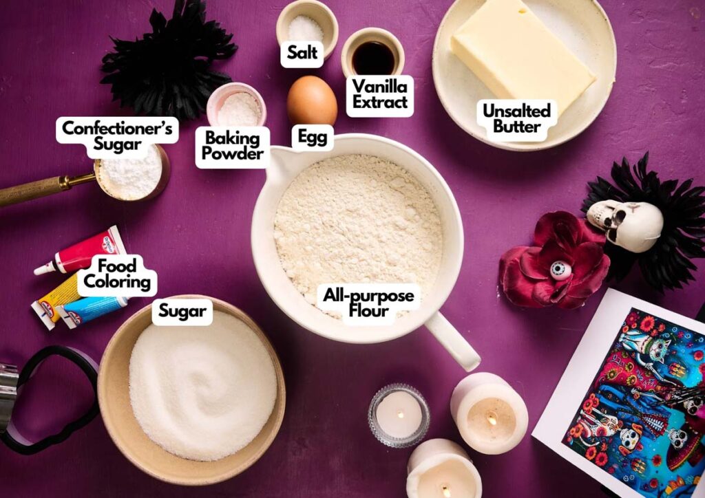Baking ingredients arranged on a purple surface for Day of the Dead Cookies, including flour, butter, egg, sugar, salt, vanilla extract, baking powder, confectioner's sugar, and food coloring.