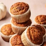 A stack of freshly baked Pumpkin Muffins in white paper liners sits on a light surface, with small pumpkins adding a festive touch in the background.