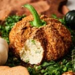 A Pumpkin-Shaped Cheeseball coated with seasoning and topped with a vegetable stem is partially sliced to reveal its creamy interior, resting on a bed of fresh herbs.