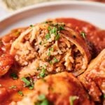 A close-up of Stuffed Cabbage Rolls topped with tomato sauce and chopped herbs on a white plate. One stuffed cabbage roll is cut open, revealing a savory filling of rice and ground meat.