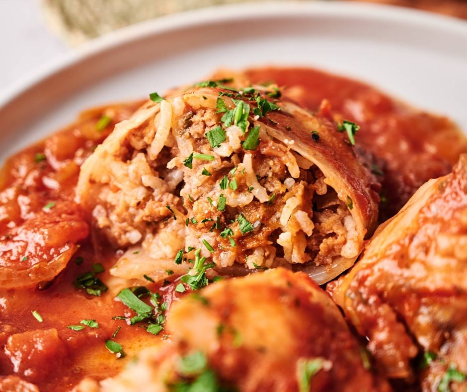 A close-up of Stuffed Cabbage Rolls topped with tomato sauce and chopped herbs on a white plate. One stuffed cabbage roll is cut open, revealing a savory filling of rice and ground meat.