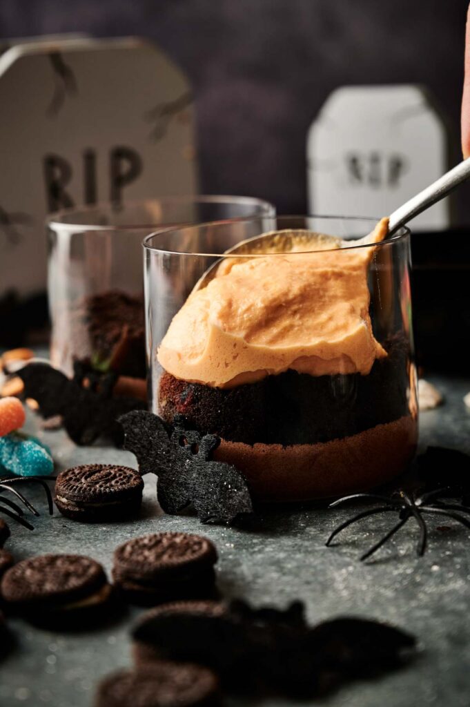 A glass dessert with layers of chocolate cake and orange cream, inspired by Halloween Dirt Cups, surrounded by cookies, candy, black bat cutouts, and festive Halloween decorations.