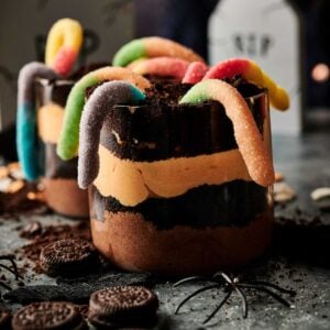Two layered chocolate and orange Halloween Dirt Cups in glass cups, topped with crushed cookies and gummy worms; Oreo cookies and a plastic spider decorate the table.