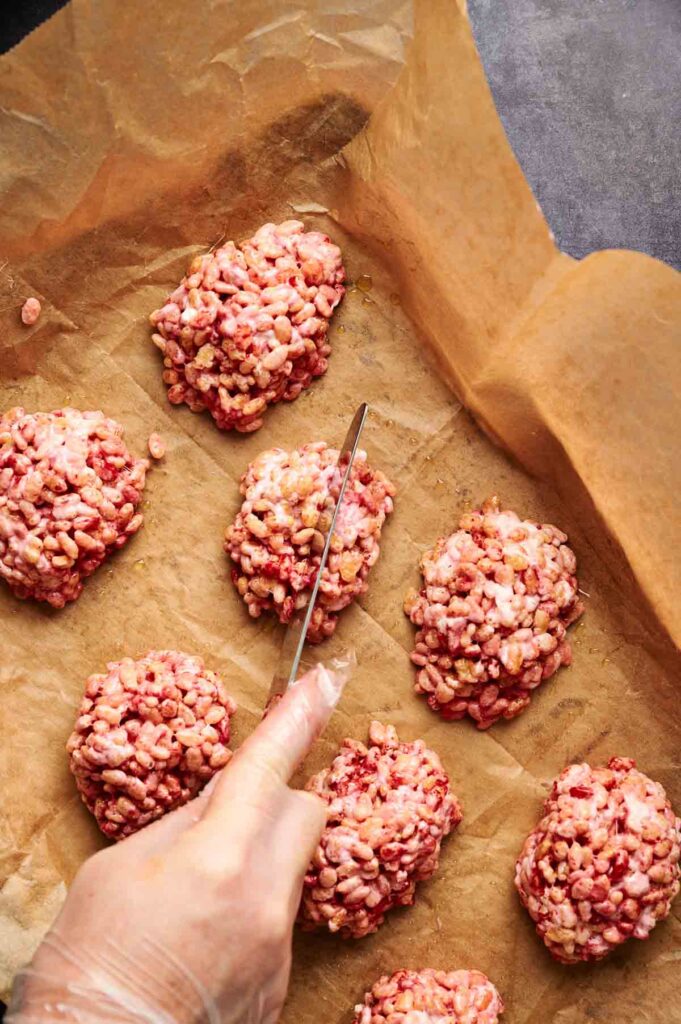 A hand wearing a glove uses a knife to cut through clusters of pink Halloween Rice Krispie Brains on a sheet of brown parchment paper.