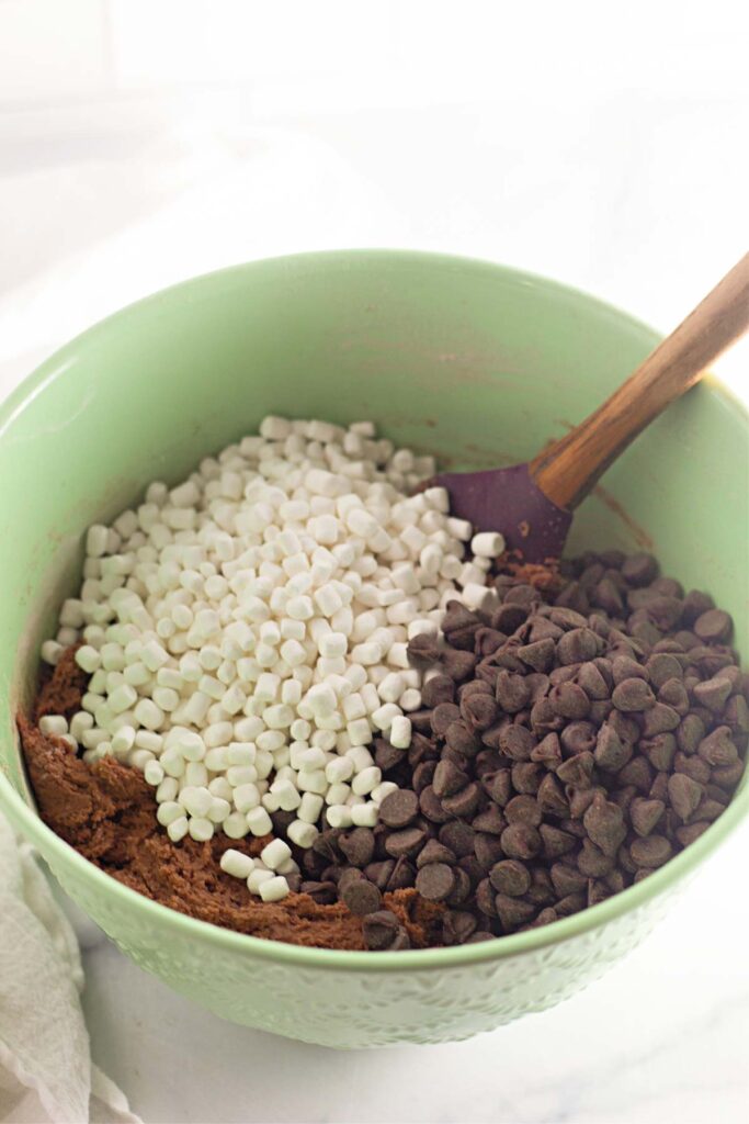 A green mixing bowl contains brownie batter, mini marshmallows, and chocolate chips—everything you need to whip up Hot Cocoa Cookies—with a spatula resting inside.