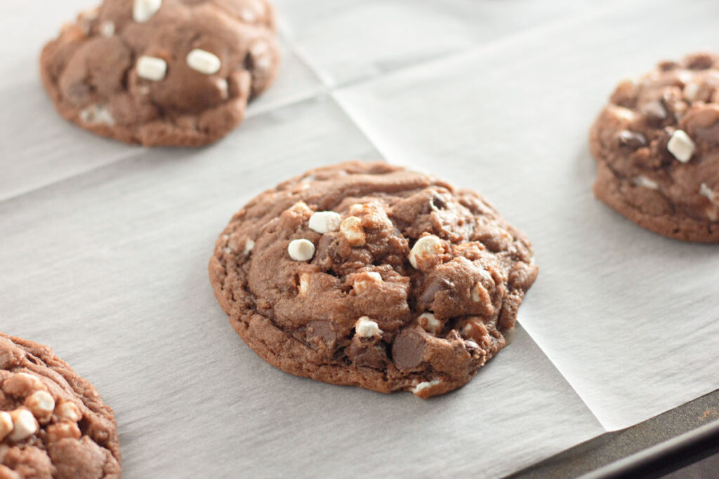 A close-up of Hot Cocoa Cookies with chocolate chips and mini marshmallows on a sheet of parchment paper.