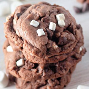 A stack of Hot Cocoa Cookies with chocolate chips and mini marshmallows sits on a light wooden surface, surrounded by scattered marshmallows and chocolate chips.