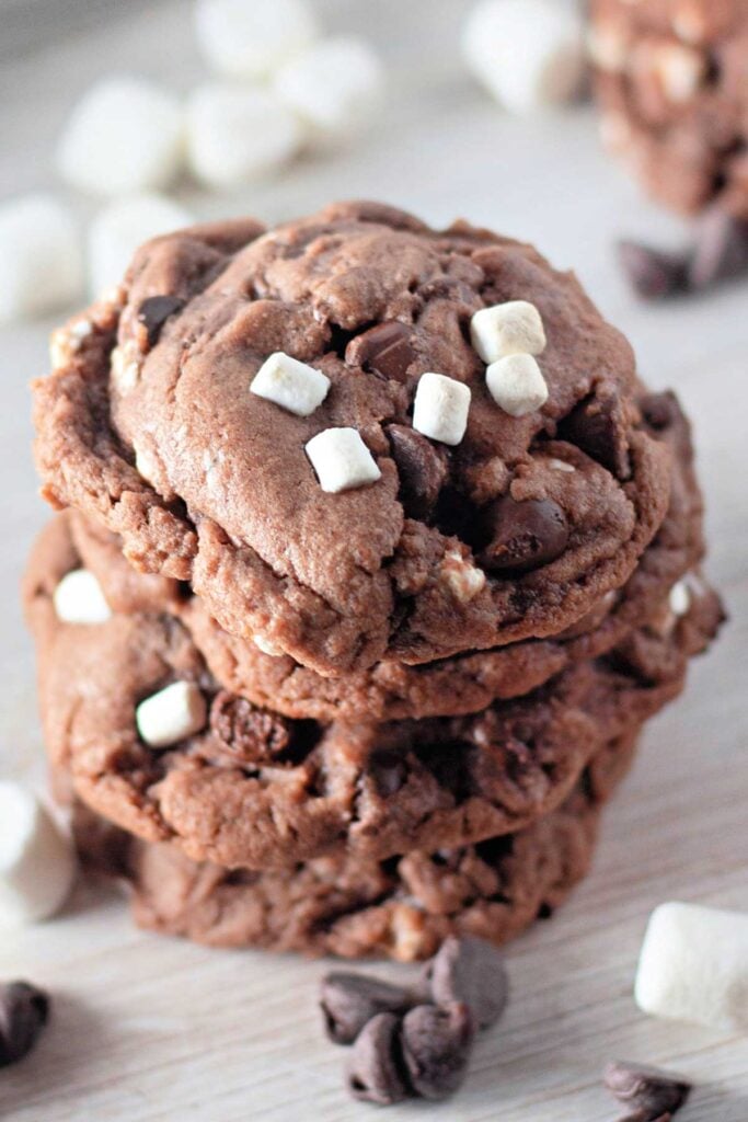A stack of Hot Cocoa Cookies with chocolate chips and mini marshmallows sits on a light wooden surface, surrounded by scattered marshmallows and chocolate chips.