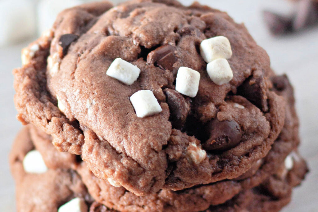 Close-up of a stack of Hot Cocoa Cookies, topped with chocolate chips and small white marshmallows for a cozy treat.