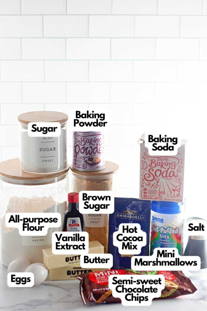 Various baking ingredients for Hot Cocoa Cookies, including flour, sugar, brown sugar, eggs, butter, vanilla extract, baking powder, baking soda, hot cocoa mix, salt, mini marshmallows, and chocolate chips.