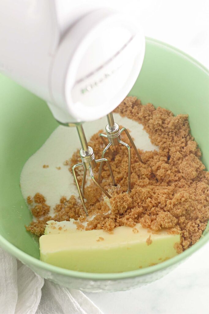 A hand mixer in a green bowl with butter, brown sugar, and white sugar being prepared for mixing delicious Hot Cocoa Cookies.