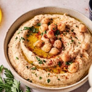 A bowl of homemade hummus topped with whole chickpeas, olive oil, paprika, and chopped parsley, with fresh parsley on the side.