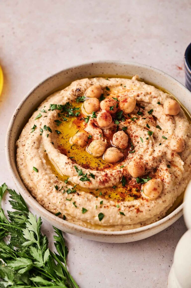 A bowl of homemade hummus topped with whole chickpeas, olive oil, paprika, and chopped parsley, with fresh parsley on the side.