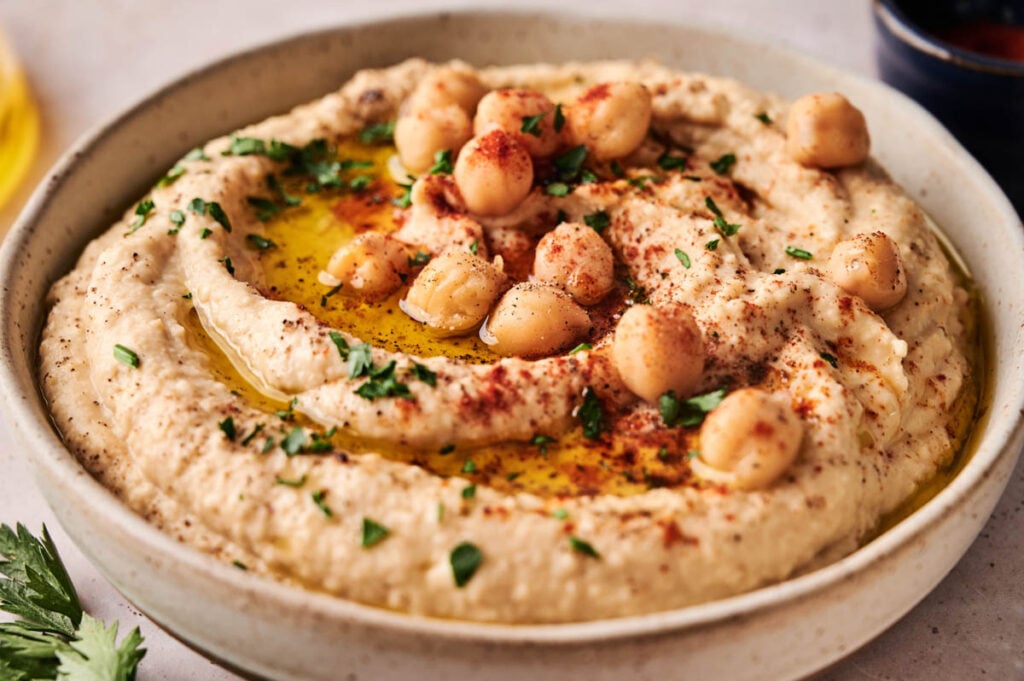 A bowl of homemade hummus garnished with whole chickpeas, olive oil, chopped parsley, and a sprinkle of paprika.
