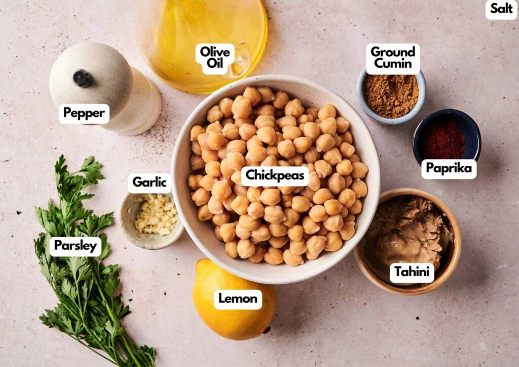Ingredients for homemade hummus, including chickpeas, tahini, lemon, garlic, parsley, pepper, olive oil, ground cumin, paprika, and salt arranged on a countertop.