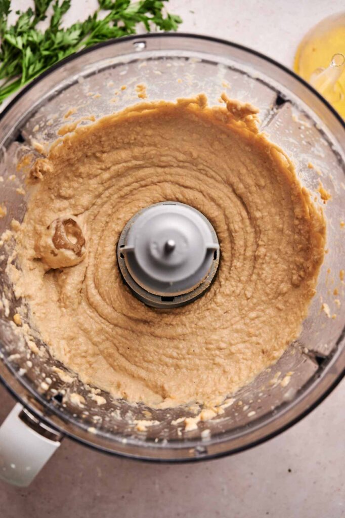 A food processor bowl filled with creamy homemade hummus, shown from above, with some unblended chickpea mixture on the sides.