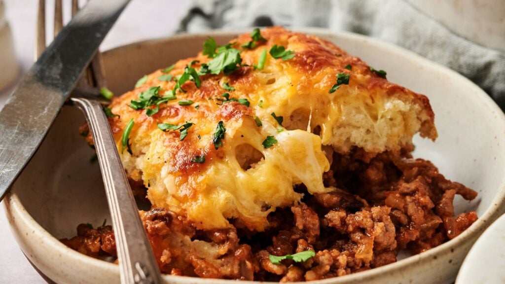 A serving of baked cheesy bread sits atop seasoned ground meat in a bowl, garnished with chopped parsley, with a fork and knife on the side.