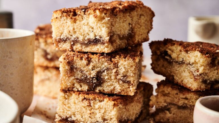 Three slices of cinnamon coffee cake are stacked on top of each other, showing a crumbly texture with a cinnamon swirl layer inside. White mugs are visible in the background.
