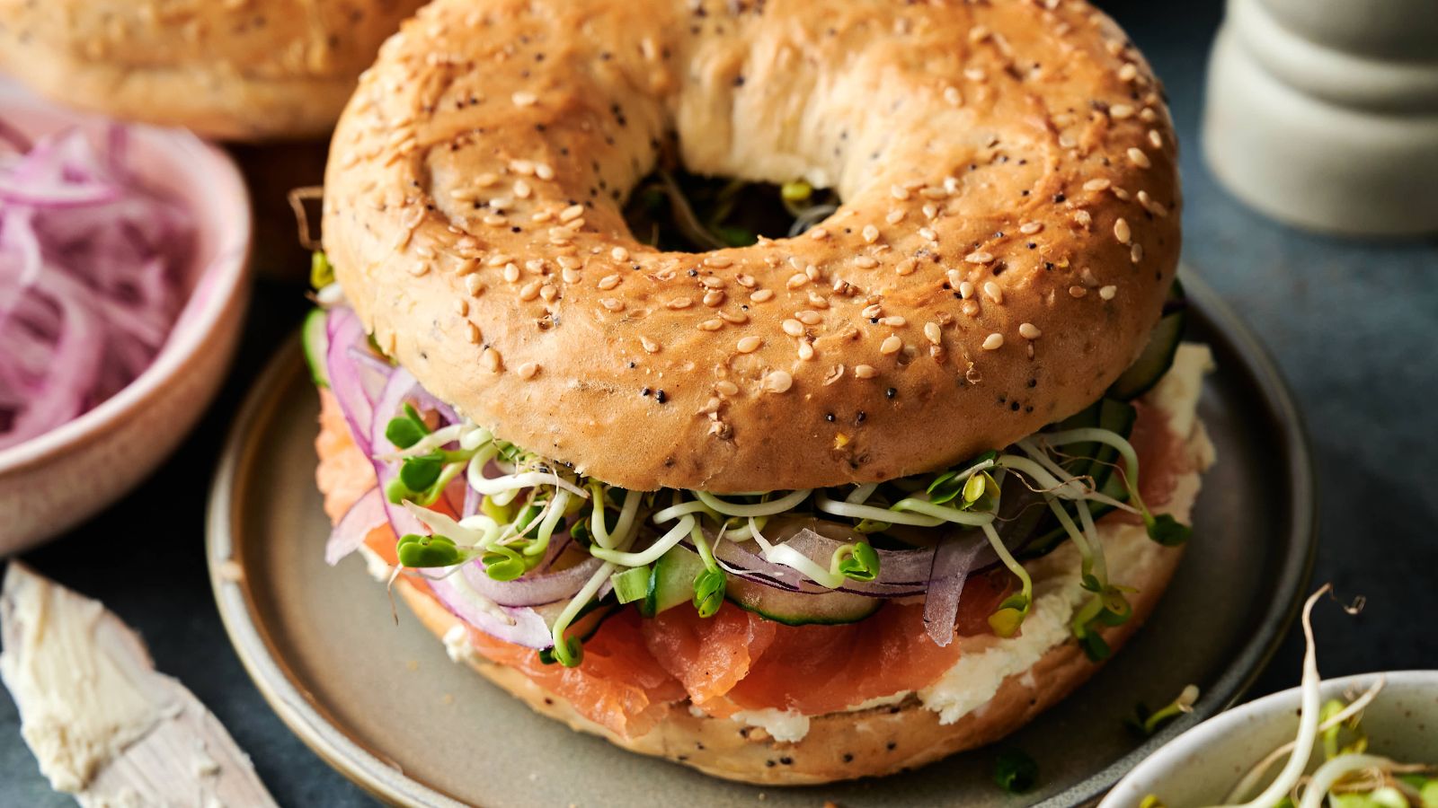 A sesame seed bagel sandwich with cream cheese, smoked salmon, red onion, cucumber, and sprouts on a plate.