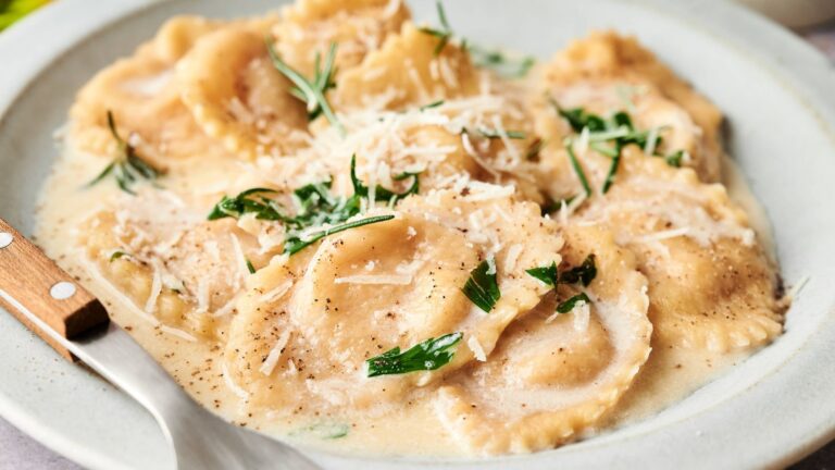 A plate of ravioli in a creamy sauce, topped with grated cheese and garnished with fresh herbs, sits next to a fork on a white plate.