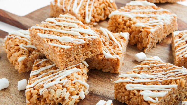 Rice crispy treats arranged on a wooden board, topped with white chocolate drizzle, with small marshmallows scattered nearby.