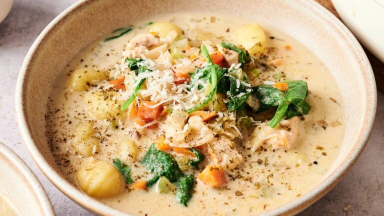 A bowl of creamy soup with gnocchi, spinach, carrots, shredded cheese, chicken, and herbs.