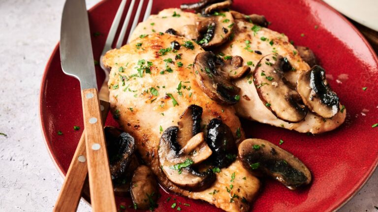 Two pieces of cooked chicken breast topped with sautéed mushrooms and herbs on a red plate, with a fork and knife placed beside them.